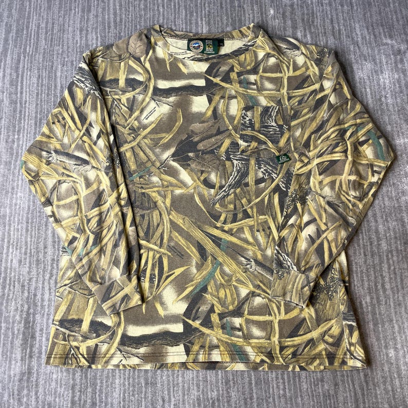 K&ouml;nnte beinhalten: Ein lang&auml;rmeliges Shirt mit Rundhalsausschnitt und einem Camouflage-Muster aus braunen, beigen und dunkelgr&uuml;nen Gr&auml;sern und Bl&auml;ttern. Das Shirt verf&uuml;gt &uuml;ber eine einzelne Brusttasche auf der linken Seite mit einem kleinen rechteckigen Markenetikett.