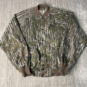 May include: A button-up jacket with a camouflage pattern in shades of green, brown, and gray. The jacket features a brown collar, cuffs, and hem, along with two chest pockets and the text "SPARTAN REALTREE".
