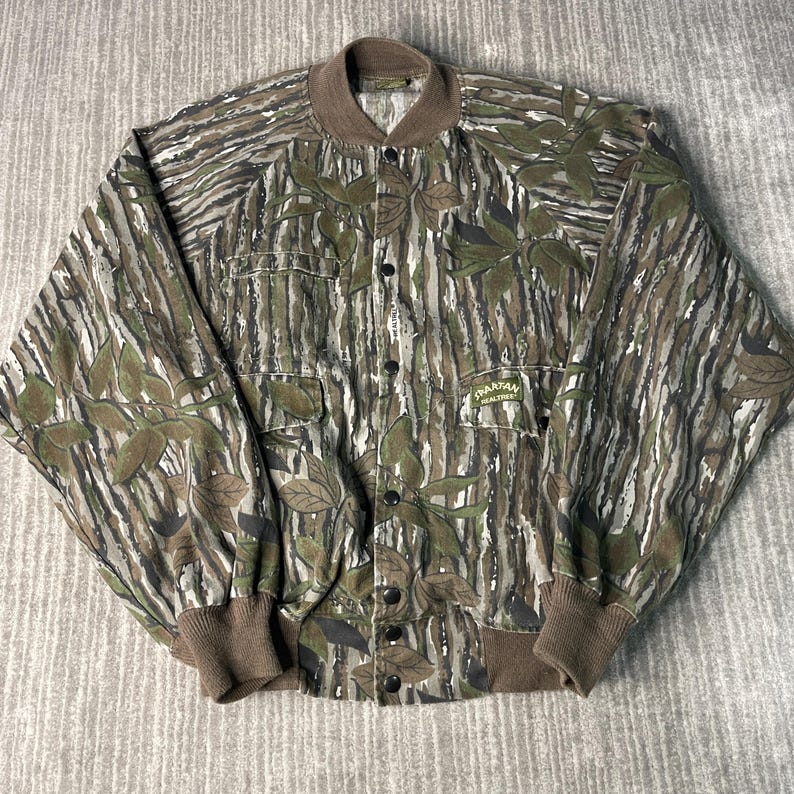 May include: A button-up jacket with a camouflage pattern in shades of green, brown, and gray. The jacket features a brown collar, cuffs, and hem, along with two chest pockets and the text "SPARTAN REALTREE".