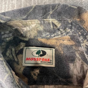 May include: Close-up of a camouflage shirt collar. The shirt features a pattern of greens, browns, and grays. A rectangular label with the text "Mossy Oak Brand Camo" is sewn onto the shirt.