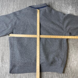 May include: A gray fleece pullover with a navy blue collar. A wooden ruler is laid across the back of the garment to measure its dimensions. The pullover has a crew neck and long sleeves. The fabric appears to be a soft, textured material.