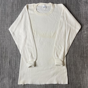 May include: A long-sleeve thermal shirt in an off-white color featuring a textured waffle-knit pattern. The garment has a crew neckline, ribbed cuffs at the wrists, and a straight hemline. A small white brand label is visible at the inner neck.