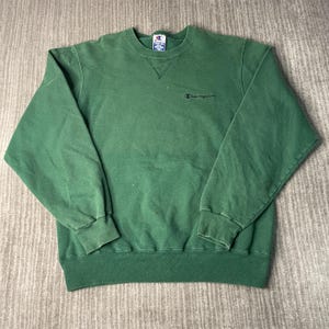May include: A green Champion sweatshirt with a crew neck and long sleeves. The Champion logo is embroidered in black on the chest. The sweatshirt has a ribbed hem and cuffs.