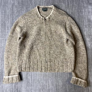 May include: A beige and brown speckled knit zip-up sweater with a round neckline and long sleeves. The garment features a ribbed hem and ribbed cuffs, with an Abercrombie & Fitch brand label visible at the inner collar. The texture is chunky.