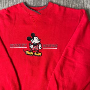 Puede incluir: Una sudadera de color rojo brillante con cuello redondo que presenta un gr&aacute;fico bordado de Mickey Mouse en el centro. El personaje aparece en negro, blanco, rojo y amarillo. El texto MICKEY MOUSE est&aacute; bordado en blanco a ambos lados.