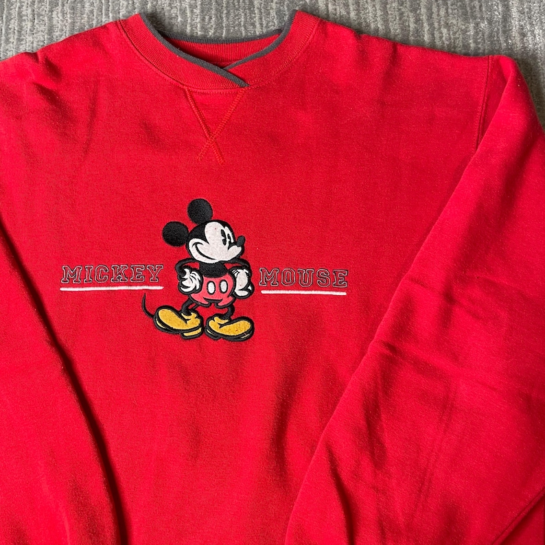 Puede incluir: Una sudadera de color rojo brillante con cuello redondo que presenta un gr&aacute;fico bordado de Mickey Mouse en el centro. El personaje aparece en negro, blanco, rojo y amarillo. El texto MICKEY MOUSE est&aacute; bordado en blanco a ambos lados.
