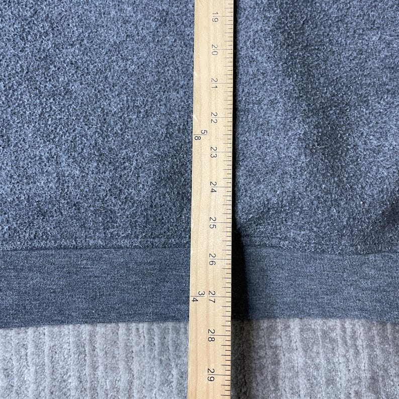 May include: A gray fabric with a dark gray band at the bottom, measured with a wooden ruler. The ruler shows measurements from 19 to 29 inches. The fabric appears to be a sweatshirt or similar garment.