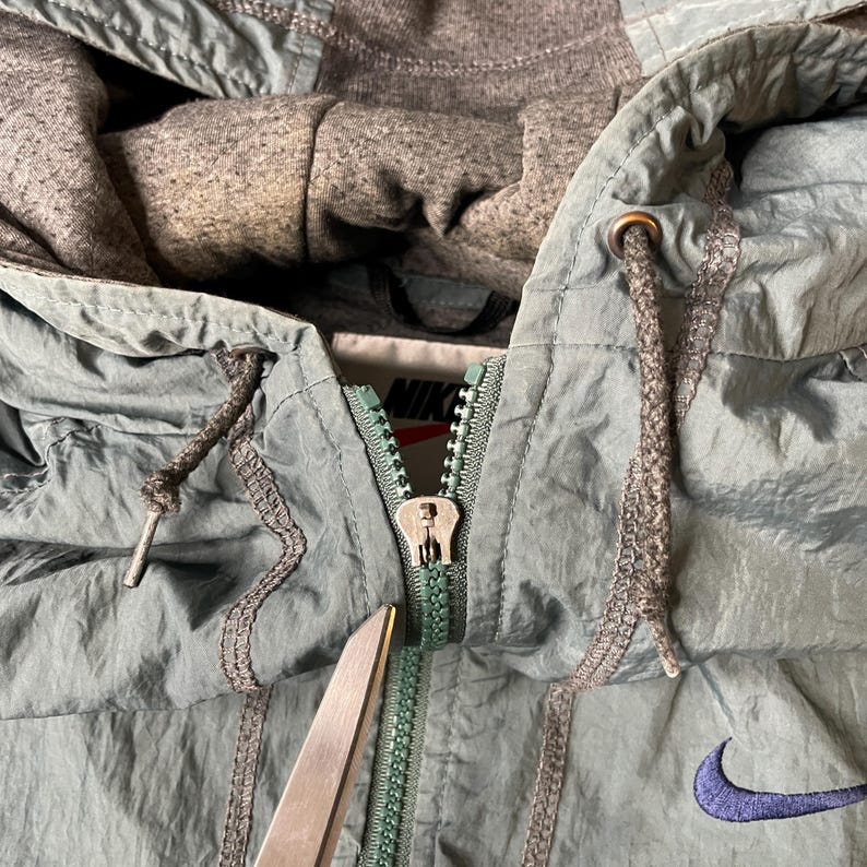 May include: A sage green hooded jacket with a crinkled texture, featuring a central zipper, a grey heathered hood lining, and a dark purple embroidered Nike swoosh logo. The interior tag displays the Nike brand name in black text on a white background.