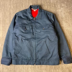 May include: A blue work jacket featuring a bright red interior lining, a pointed collar, and a full-length front zipper. The garment includes two chest pockets with buttoned flaps and two slanted side pockets, finished with buttoned cuffs.