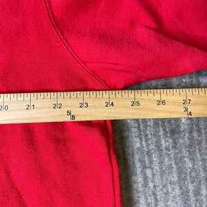 Puede incluir: Una prenda de color rojo brillante con una costura visible a lo largo del hombro y la manga. Una regla de madera est&aacute; colocada horizontalmente sobre la tela, mostrando marcas de medici&oacute;n de 50,8 a 68,6 cm con indicadores fraccionarios.