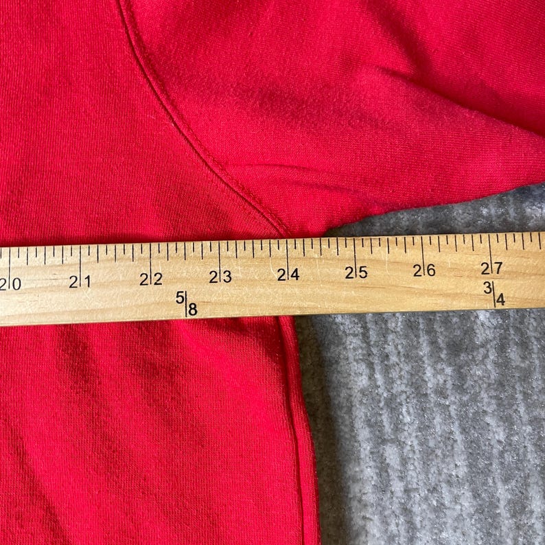 Puede incluir: Una prenda de color rojo brillante con una costura visible a lo largo del hombro y la manga. Una regla de madera est&aacute; colocada horizontalmente sobre la tela, mostrando marcas de medici&oacute;n de 50,8 a 68,6 cm con indicadores fraccionarios.