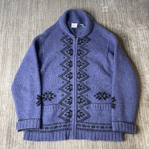 May include: A periwinkle blue zip-up sweater with a shawl collar and black geometric patterns. The sweater has two front pockets and ribbed cuffs and hem. The zipper is silver. The sweater is made of a thick knit material.