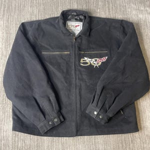 Puede incluir: Una chaqueta negra de manga larga con cremallera frontal completa, cuello y dos bolsillos con cremallera en el pecho. El pecho presenta un logotipo bordado del 50 aniversario de Corvette con una bandera a cuadros y un emblema rojo.