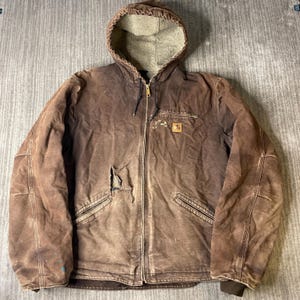 May include: A brown hooded jacket with a beige fleece lining, a full-length front zipper, and a small square logo patch on the chest. The jacket features two lower pockets, a zippered chest pocket, and a visible tear on the front right side.