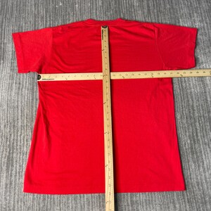May include: A vibrant red t-shirt with short sleeves, displayed flat on a grey surface. A wooden ruler is positioned across the shirt, measuring its dimensions. The shirt's material looks soft and slightly worn.