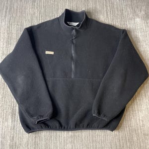 May include: A black fleece pullover featuring a mock neck collar and a partial front zipper. The garment includes long sleeves with elastic cuffs, a straight hem, and a small rectangular tan Columbia brand patch on the upper right chest area.