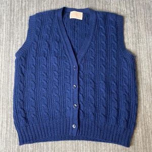 May include: A navy blue sleeveless knit sweater vest featuring a V-neckline, a vertical cable knit pattern, and a front button closure with four matching buttons. The garment includes a ribbed hem and a Pendleton brand label at the inner neckline.