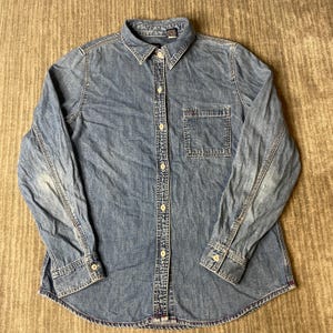 May include: A long-sleeve button-down shirt made of blue denim fabric. The shirt features a standard collar, a single chest pocket on the left side, and light-colored buttons running down the front placket and on the sleeve cuffs.
