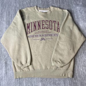 May include: A tan crewneck sweatshirt featuring maroon and white text that reads MINNESOTA, ESTABLISHED EIGHTEEN FIFTY-ONE, GOLDEN GOPHERS, TWIN CITIES, and MINNESOTA, along with a maroon M logo. The long-sleeve garment has a ribbed collar and cuffs.