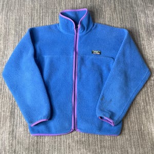 May include: A blue fleece jacket with a full zip and a high collar. The jacket features purple trim on the collar, cuffs, and zip. A small rectangular patch is on the chest. The jacket is made of a soft, textured material.