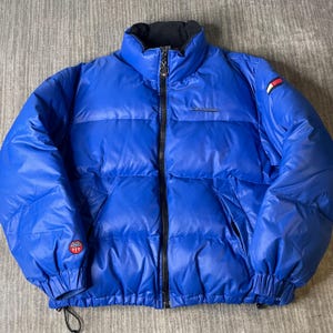 May include: A vibrant blue puffer jacket with a black zipper and collar. The jacket features a small logo on the sleeve and a small logo on the chest. The jacket has a drawstring at the bottom and elastic cuffs.