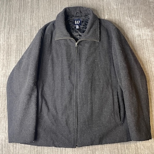 May include: A charcoal grey zip-up jacket with a fold-down collar and long sleeves. The jacket features a black quilted interior lining and a central zip closure. A black label with the white text GAP is visible at the inner neckline.