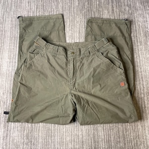 May include: Olive green outdoor pants with multiple pockets and a button closure. The pants have a drawstring at the ankles and a small red logo patch on the right side. The pants are made of a lightweight, durable material.