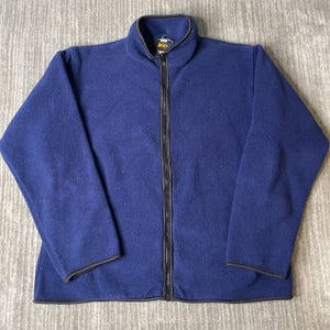 May include: A solid navy blue fleece jacket featuring a full-length front zipper, a high mock neck collar, and dark trim along the edges of the collar, cuffs, and hem. The interior neck label displays the REI brand logo in yellow and white text.