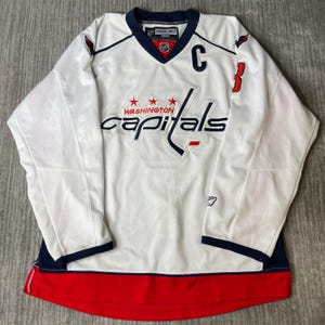 May include: A white Washington Capitals hockey jersey with navy blue and red accents. The front features the team name in navy blue text with three red stars above it, a navy blue captain's C, and a red hem. The Reebok logo is visible.