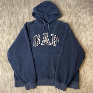 Könnte beinhalten: Ein marineblauer Fleece-Kapuzenpullover mit dem Schriftzug GAP in großen weißen Blockbuchstaben mit dünner marineblauer Umrandung und dem Wort Athletic in kleinerer weißer Schreibschrift unter dem GAP-Logo sowie einer Kängurutasche.