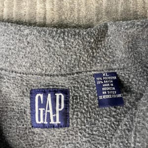 May include: Close-up of a gray fleece jacket with a navy blue GAP label. The jacket has a tag with the size XL and fabric composition details. The tag also indicates the jacket was made in Indonesia.