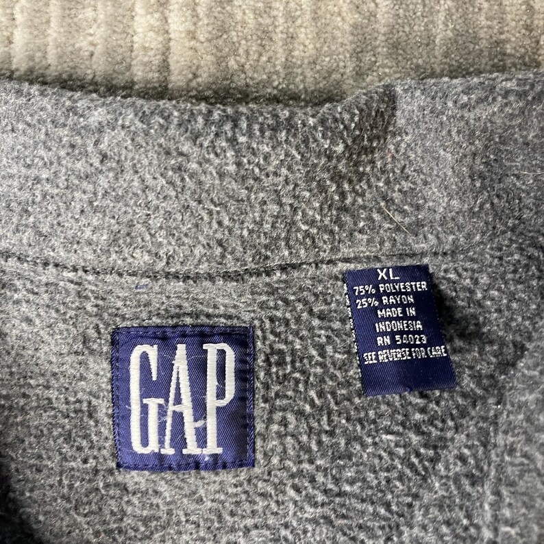 May include: Close-up of a gray fleece jacket with a navy blue GAP label. The jacket has a tag with the size XL and fabric composition details. The tag also indicates the jacket was made in Indonesia.