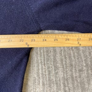 Puede incluir: Una prenda de punto azul oscuro con textura acanalada, colocada detr&aacute;s de una regla de madera que muestra medidas de 50,8 a 71,1 cm, indicando claramente el ancho de la secci&oacute;n de tela en el &aacute;rea del pecho de la prenda.