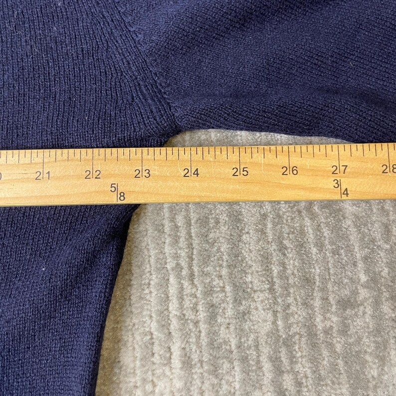 Puede incluir: Una prenda de punto azul oscuro con textura acanalada, colocada detr&aacute;s de una regla de madera que muestra medidas de 50,8 a 71,1 cm, indicando claramente el ancho de la secci&oacute;n de tela en el &aacute;rea del pecho de la prenda.