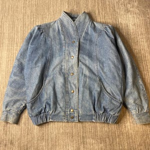 May include: A light blue denim jacket featuring a high collar, long sleeves with gathered shoulders, and a front placket with five metallic snap buttons. The jacket includes two side pockets and an elasticized hemline at the waist.