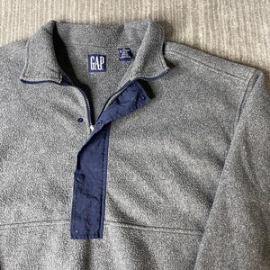 May include: A gray fleece pullover with a navy blue accent on the placket and collar. The GAP logo is visible on the chest. The pullover has a zipper and snap closure.