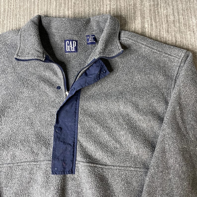 May include: A gray fleece pullover with a navy blue accent on the placket and collar. The GAP logo is visible on the chest. The pullover has a zipper and snap closure.