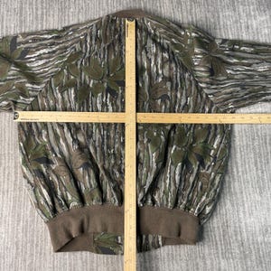 May include: A camouflage bomber jacket with a brown collar and cuffs. The jacket features a pattern of green leaves and brown and gray stripes. A wooden ruler is laid across the jacket.