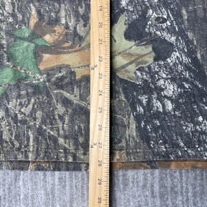 May include: A camouflage-patterned t-shirt with a ruler showing measurements in inches. The shirt features a mix of green, brown, and black colors, mimicking a forest environment. The bottom of the shirt has the text "BY OAK".