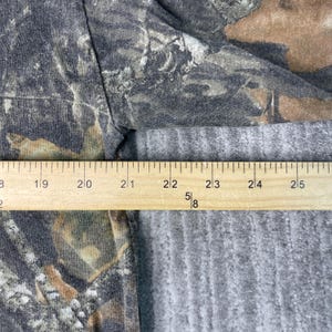May include: A close-up of a camouflage pattern fabric with a wooden ruler. The ruler shows measurements in inches, with markings from 18 to 25 inches. The fabric has shades of green, brown, and gray.
