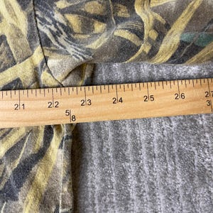 K&ouml;nnte beinhalten: Ein Kleidungsst&uuml;ck mit einem gelben, braunen und schwarzen Tarnmuster. Ein Holzlineal liegt horizontal auf dem Stoff, wobei die Messmarkierungen deutlich sichtbar sind und eine Breite von 52 bis 69 Zentimetern anzeigen.