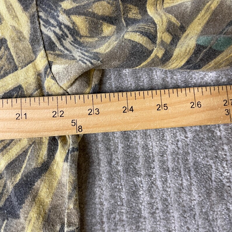 K&ouml;nnte beinhalten: Ein Kleidungsst&uuml;ck mit einem gelben, braunen und schwarzen Tarnmuster. Ein Holzlineal liegt horizontal auf dem Stoff, wobei die Messmarkierungen deutlich sichtbar sind und eine Breite von 52 bis 69 Zentimetern anzeigen.