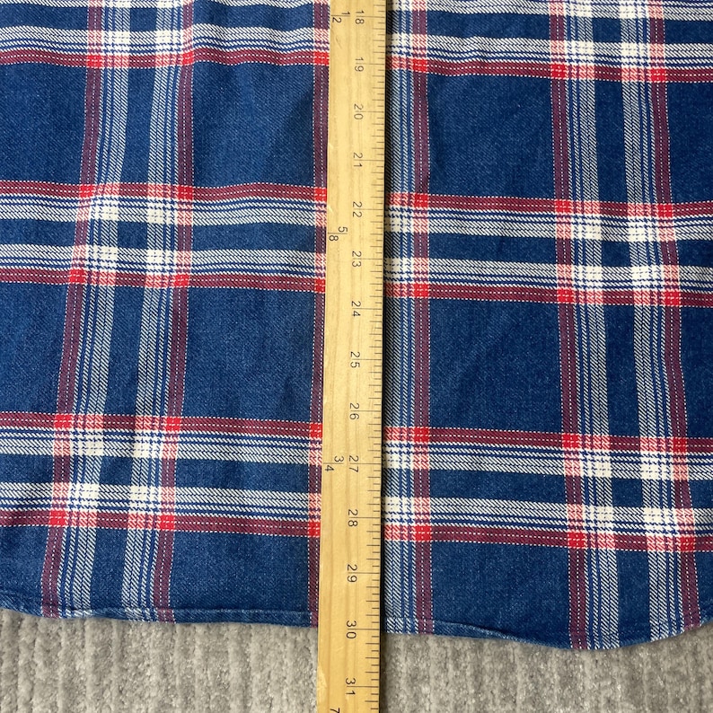 May include: Close-up of a blue, red, and white plaid fabric. The fabric is measured with a wooden ruler, showing measurements from 18 to 31 inches. The plaid pattern features thick blue lines with red and white accents.