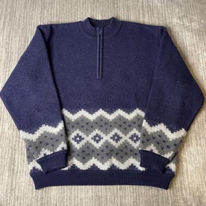 May include: A navy blue quarter-zip sweater featuring a textured knit fabric. The lower portion of the garment and the sleeves are decorated with a repeating geometric pattern in shades of light gray and dark gray, finished with ribbed cuffs.
