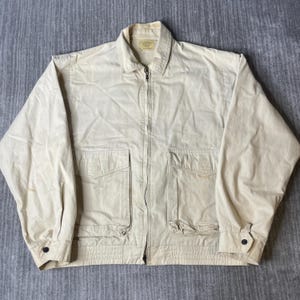 May include: A beige zip-up jacket featuring a classic collar, long sleeves with buttoned cuffs, and two large front patch pockets with flaps. The garment has a structured fit with a ribbed hem and a central metal zipper closure extending to the neck.