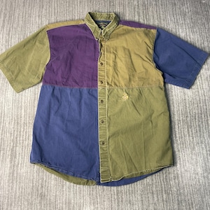 May include: A short-sleeved button-down shirt with a color-block design. The shirt features panels of olive green, purple, and blue. A small embroidered emblem is on the front. The shirt has a button-down collar and a relaxed fit.