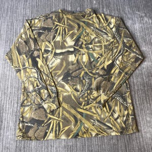 K&ouml;nnte beinhalten: Ein lang&auml;rmeliges Shirt mit Rundhalsausschnitt und einem Tarnmuster in Braun-, Beige- und Dunkelgr&uuml;nt&ouml;nen. Der Aufdruck zeigt einen dichten Waldboden mit verstreuten Bl&auml;ttern, Zweigen und &Auml;sten, die das gesamte Kleidungsst&uuml;ck vom Kragen bis zum Saum bedecken.