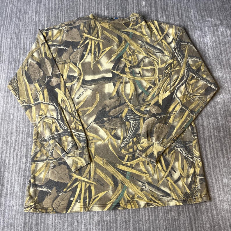 K&ouml;nnte beinhalten: Ein lang&auml;rmeliges Shirt mit Rundhalsausschnitt und einem Tarnmuster in Braun-, Beige- und Dunkelgr&uuml;nt&ouml;nen. Der Aufdruck zeigt einen dichten Waldboden mit verstreuten Bl&auml;ttern, Zweigen und &Auml;sten, die das gesamte Kleidungsst&uuml;ck vom Kragen bis zum Saum bedecken.