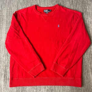 May include: A bright red long-sleeve crewneck sweatshirt featuring a small blue embroidered pony logo on the left chest and a dark blue Polo by Ralph Lauren brand label at the inner neckline. The garment has ribbed cuffs and a ribbed hem.