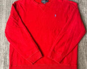 May include: A bright red long-sleeve crewneck sweatshirt featuring a small blue embroidered pony logo on the left chest and a dark blue Polo by Ralph Lauren brand label at the inner neckline. The garment has ribbed cuffs and a ribbed hem.